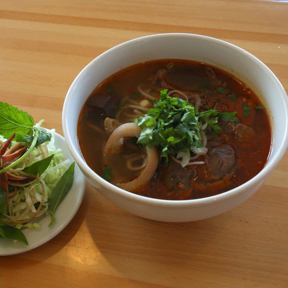 A bowl of Vietnamese Bun Bo Hue soup with a side plate of fresh herbs and vegetables.