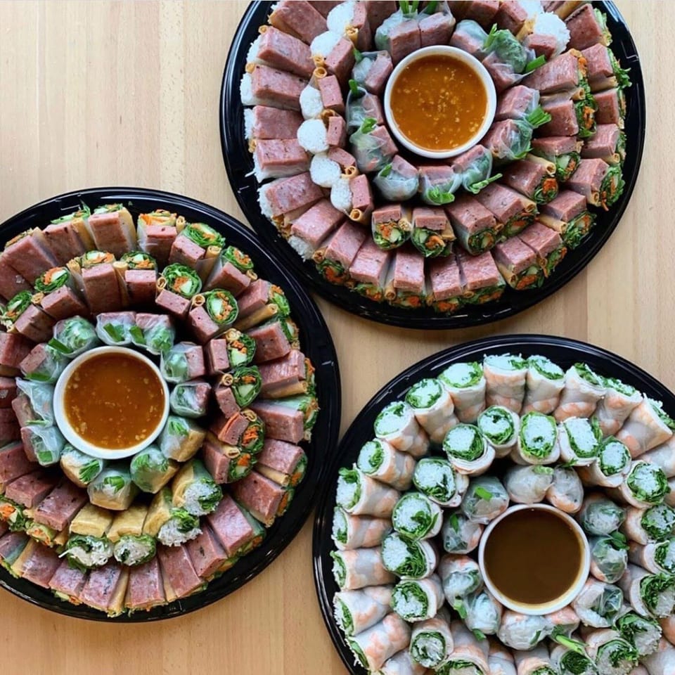 Three platters of Vietnamese spring rolls and spam musubi, with dipping sauces, on a wooden table.