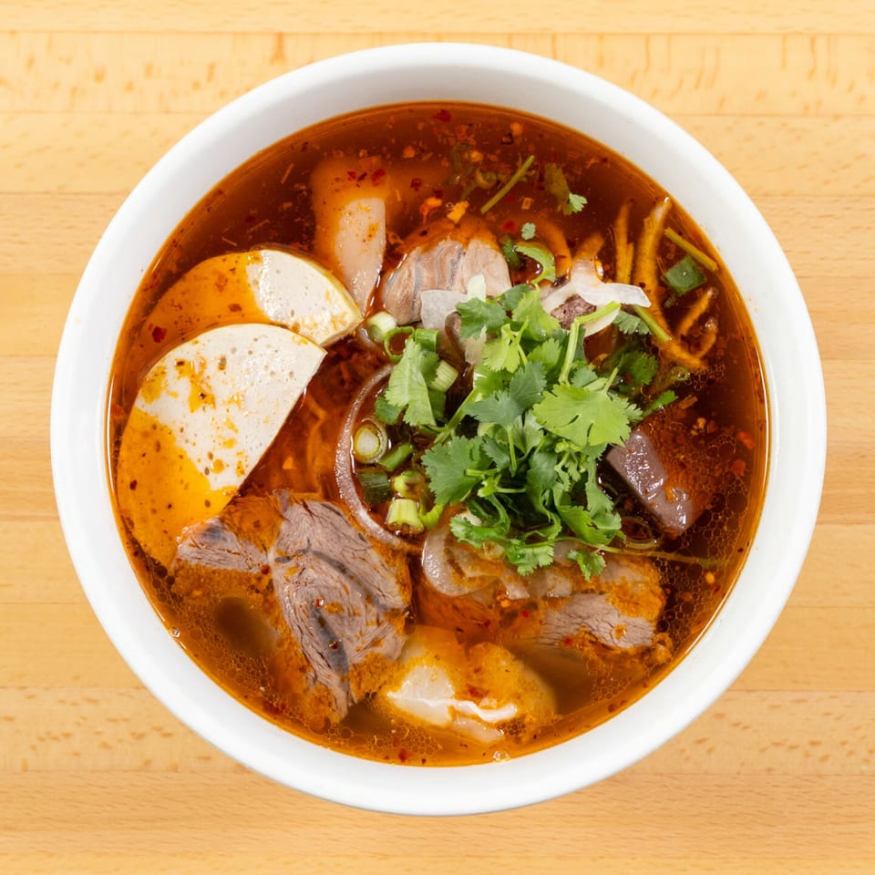 Overhead view of a vibrant Bún Bò Huế soup with beef, pork, and herbs in a white bowl.